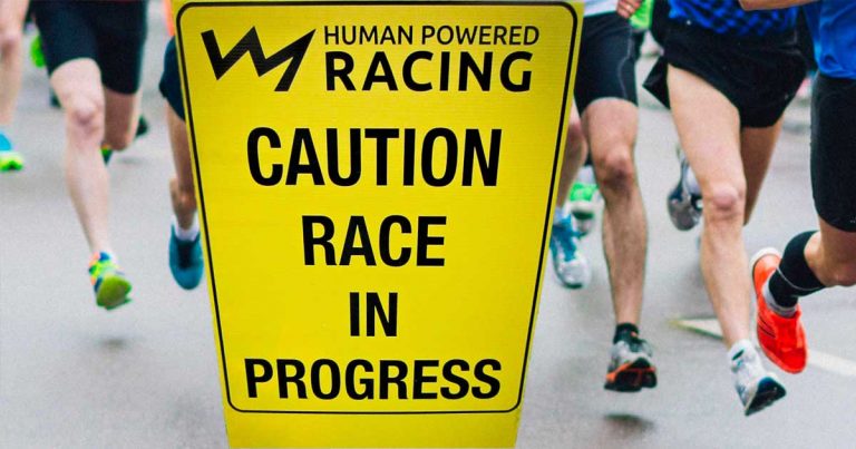 Runners run by the Human Powered Racing sign: caution race in progress.