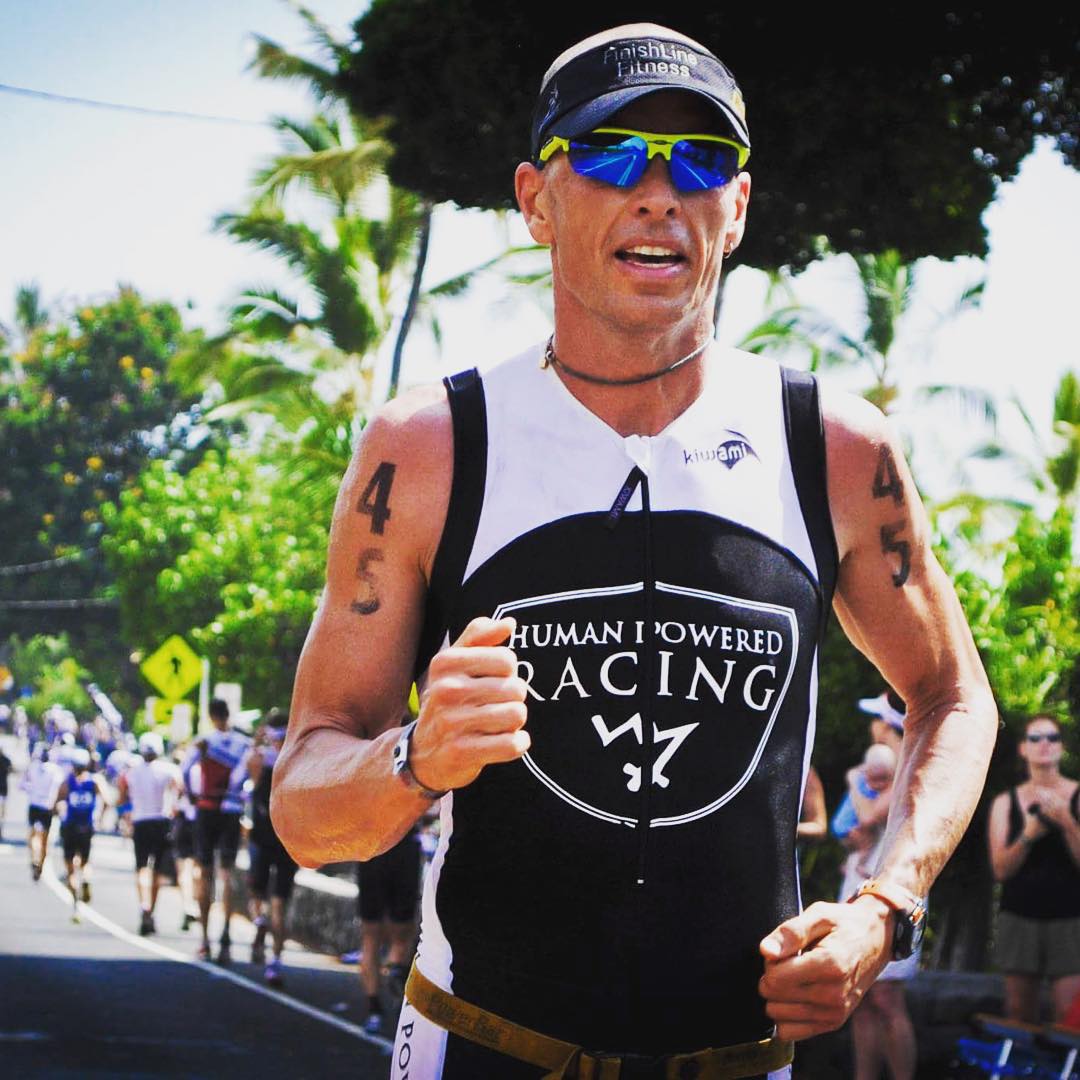 A man running in a white and black triathlon body suit. We can see from his hips upwards. Across his chest it says "Human Powered Racing." He is also wearing a black visor and blue sunglasses. He is running, and there is a crowd of runners and a blue sky and some palm trees visible behind him.