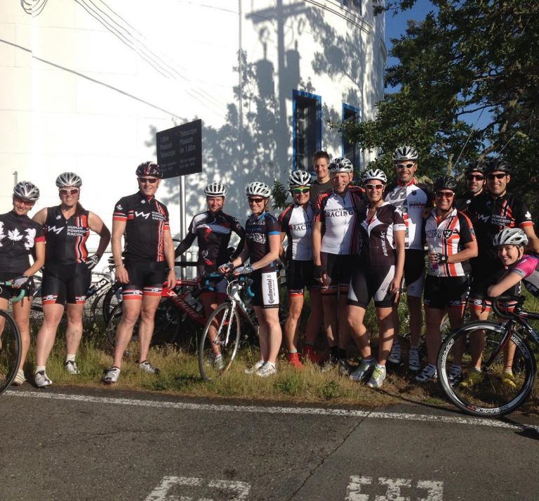 A group of bikers with their bikes standing outside a white building on a sunny day. Beside the building on the right the branches of a green tree come into the photo and a between them a blue sky is visible. The bikers are smiling, and in full biking gear.