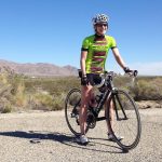 A woman in a lime green tshirt and biking shorts with a helmet on a bike. Her feet are on the ground stabilizing the bike, and the landscape around her is dry with gravel sparse bush, brown mountains in the distance, and a blue sky.