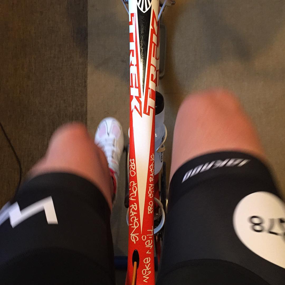 Taken looking downwards at someone's legs while they are sitting on a red, white, and black bicycle. Only the middle bar of the bike and the person's thighs, knees, and left foot are visible. They are wearing black bike shorts with the white Human Powered Racing logo on the left, which is a zig zag line shaped like the silhouette of a mountain range.