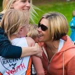 Two adult women celebrating with a young girl. All are blonde, and the two woman are smiling widely. We cannot see the girl's face. The woman behind the girl has her right arm around the girl and is squeezing her shoulder. The other woman is kneeling down and is face to face with the young girl and smiling. The grass in the background is bright green.