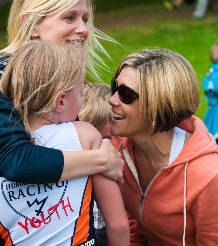 Two adult women celebrating with a young girl. All are blonde, and the two woman are smiling widely. We cannot see the girl's face. The woman behind the girl has her right arm around the girl and is squeezing her shoulder. The other woman is kneeling down and is face to face with the young girl and smiling. The grass in the background is bright green.