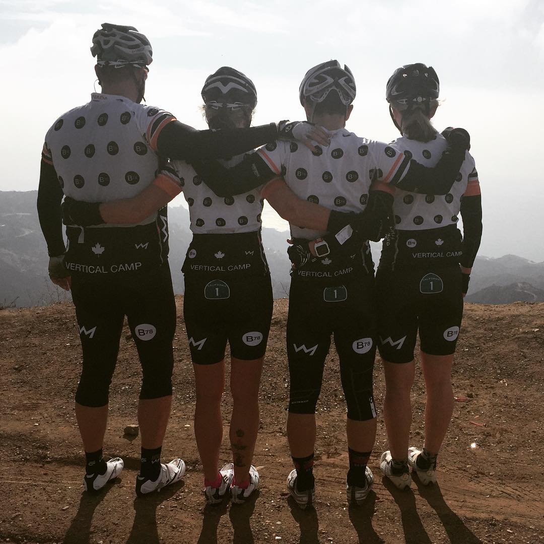 Four people from behind. They all have their arms around each other, and are wearing matching biking gear. They are wearing black shorts and white tshirts with black polkadots, helmets, and bike shoes. The view in front of them is hills and fog. They are standing on gravel.