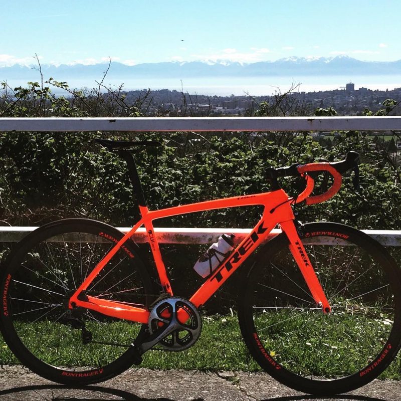 A bright red bike with "TREK" printed in bold black letters on the lower bar. It is leaning against a two rung metal railing, and there are leafy trees, mountains, and light blue sky in the background.