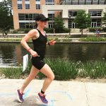 A woman in a black tank and shorts with a black baseball cap. She is running and behind her is a pond and a sandy coloured beige and cream coloured building in the background.