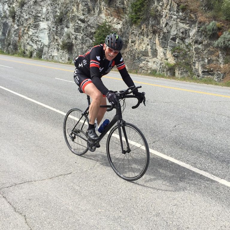 A man biking up a slight hill on a road. He is looking to his right towards the camera and smiling.