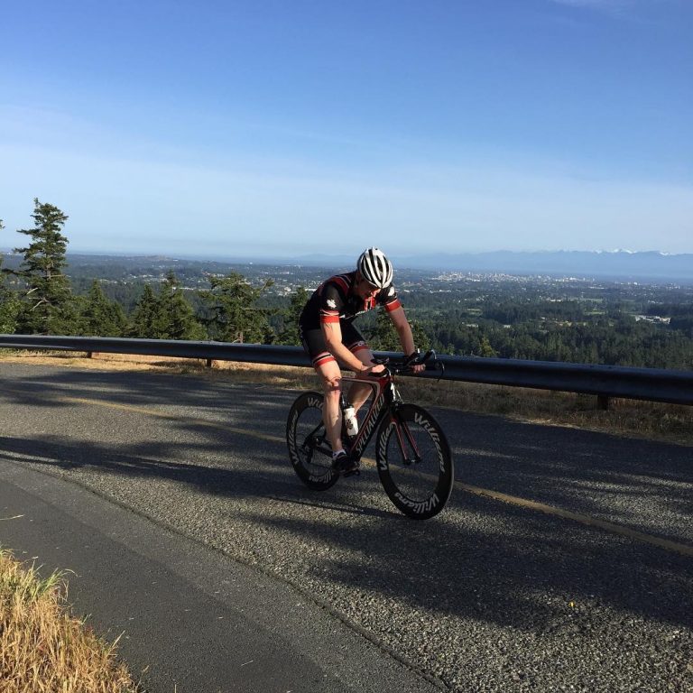 A man on a road biking: he is in the middle of the road and the photo is taken from the left. Past the road is a green city scape and a blue sky.