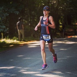 A woman running towards the camera on a road with forest in the background. She is wearing a black triathlon suit with a red strip down her left side. She has a race number pinned to her lower torso.