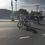 Two bikers on a road with a low metal fence, green trees, and blue cloudy sky in the distance. One biker in the front and centre is closest to the camera and the focus of the photo. They other is behind and further back.