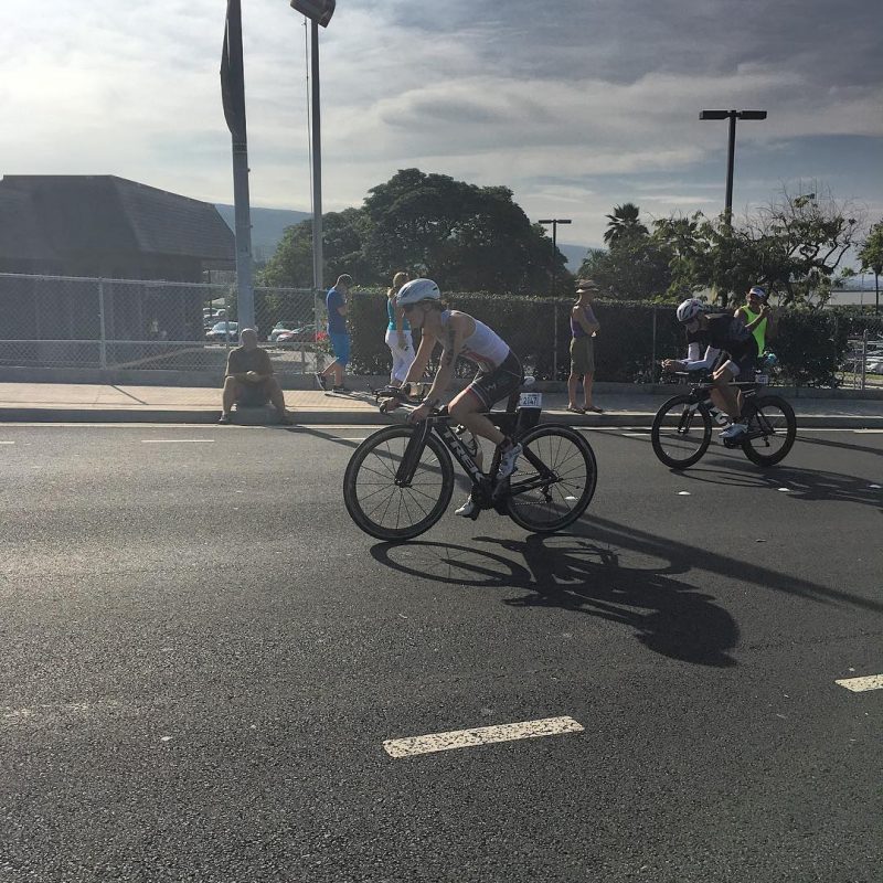 Two bikers on a road with a low metal fence, green trees, and blue cloudy sky in the distance. One biker in the front and centre is closest to the camera and the focus of the photo. They other is behind and further back.