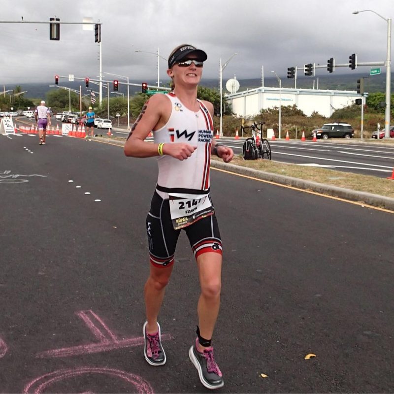 A woman running towards us on a road. She is wearing a triathlon bodysuit in the HPR colours of red, white, and black. She is also wearing a black visor. It is a grey, cloudy day, and there is a four way stop cluttered with orange directional cones in the background