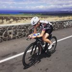 A woman leaning over the handlebars of he black bicycle as she bikes along a paved road. There is a low stone wall to her right, and beyond that a dry grassy area, and a deep blue sky that gets cloudy and brighter higher up.