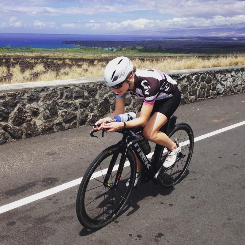 A woman leaning over the handlebars of he black bicycle as she bikes along a paved road. There is a low stone wall to her right, and beyond that a dry grassy area, and a deep blue sky that gets cloudy and brighter higher up.