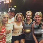 A group of five people shoulder to shoulder outside at night. All of them are smiling, and there is some light coming from the upper left. The woman second from the right is holding a wooden bowl with a plaque on it.