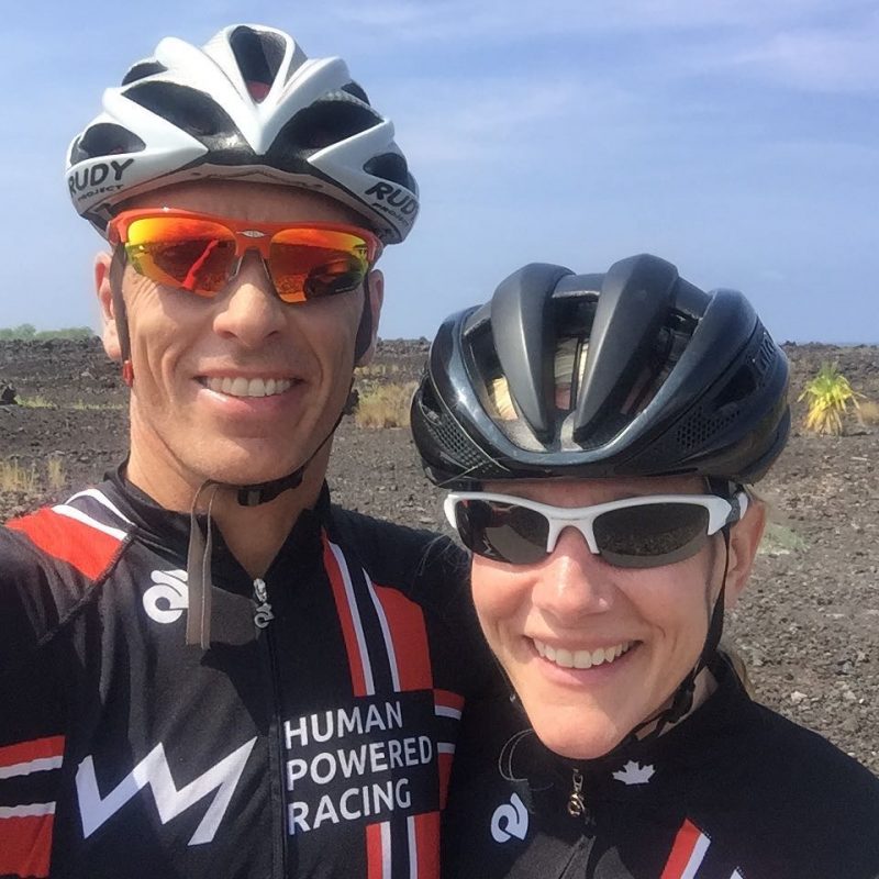 A man and a woman from the chest up with the man on the left and the woman on the right. Both are smiling with HPR black and red tops, sunglasses, and helmets. The background is a dry grassy field and a blue sky.