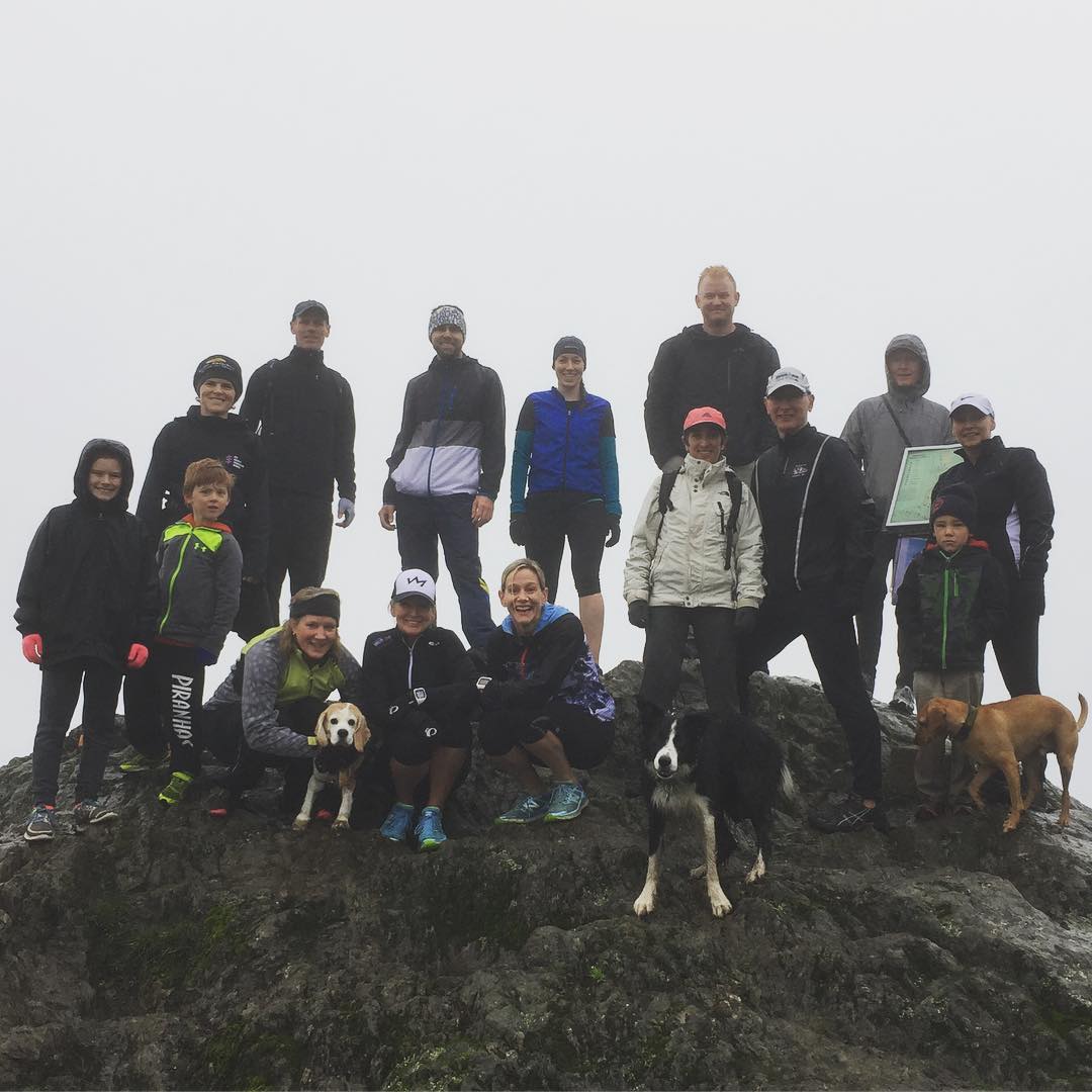 A group of people in winter outdoors gear and three dogs standing at the top of a rocky point against a backdrop of thick fog.