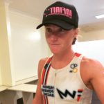 A woman's torso and face taken slightly to her left. She is wearing a black cap, and is sweaty and flushed. She has a red and white HPR tank and her lips are slightly drawn in. She looks focussed.