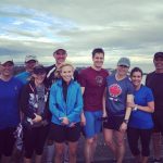 Group of people in running gear. They are outside on a cloudy day, and the city scape of victoria is just visible behind them. From their hair and clothes it looks windy.