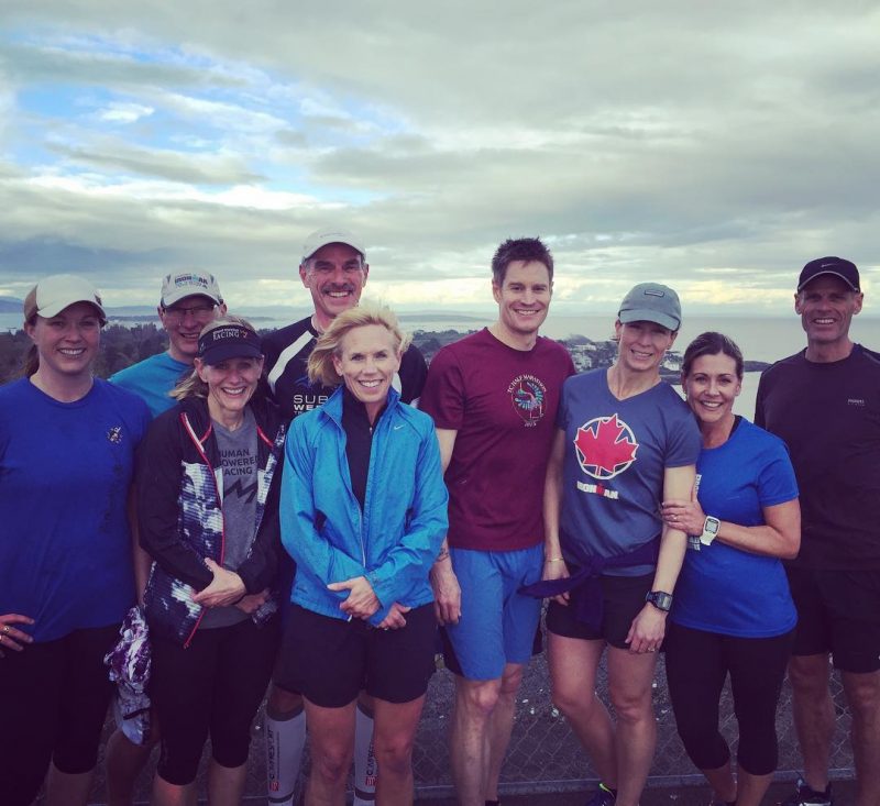 Group of people in running gear. They are outside on a cloudy day, and the city scape of victoria is just visible behind them. From their hair and clothes it looks windy.