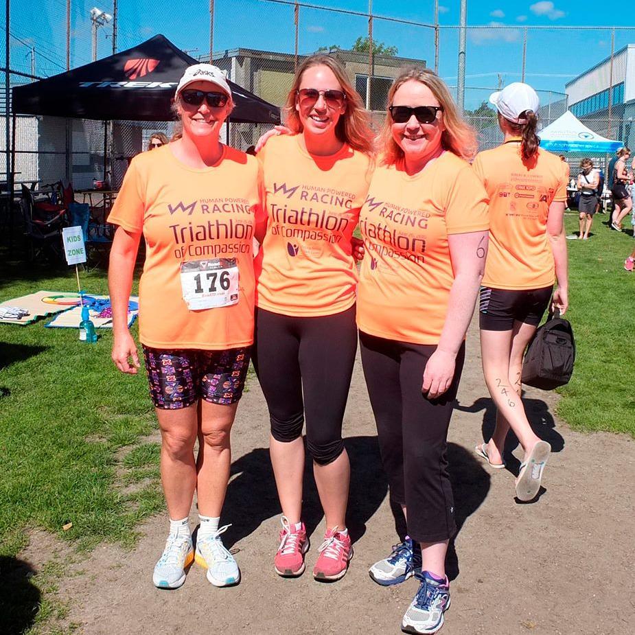 Three women standing shoulder to shoulder with their arms around each other. There are wearing matching orange shirts that say "Human Powered Racing Triathlon of Compassion" and are standing on a grassy green field.