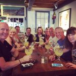 A group sitting around a long wooden table. It is a large group of about nine people, and they all have yellow margaritas in margarita glasses. All are smiling at the camera, which is positioned at the front of the table.
