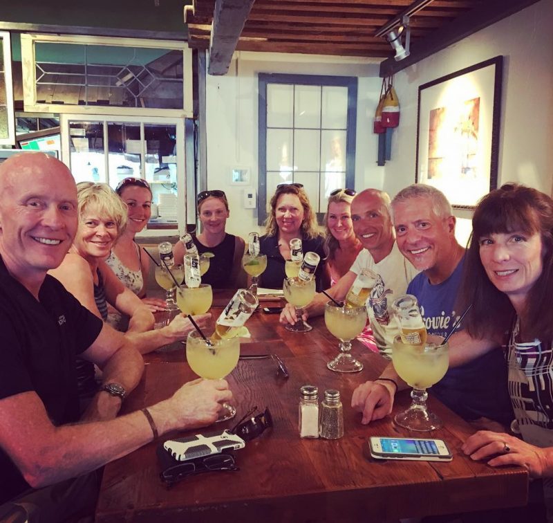 A group sitting around a long wooden table. It is a large group of about nine people, and they all have yellow margaritas in margarita glasses. All are smiling at the camera, which is positioned at the front of the table.