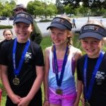 Three young girls with medals on blue ribbons around their necks are standing shoulder to shoulder and smiling. They are all wearing running clothes and matching black "Human Powered Racing" visors. The back ground looks like field, and then a paved area, and then a patch of trees on a grey, but slightly sunny day. The background it out of focus.