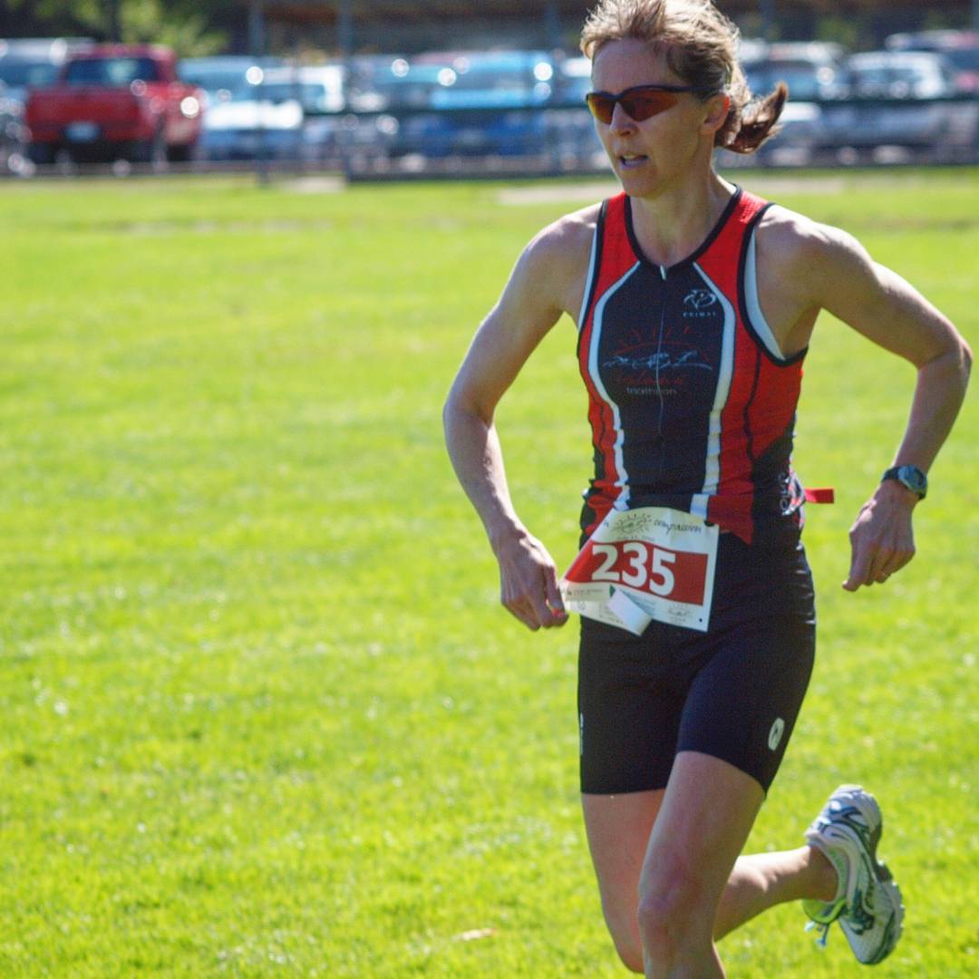 A woman running in a red, white, and black HPR triathlon bodysuit with sunglasses. Her race number is pinned to her stomach and there is a green field and then a parking lot in the background. It is sunny.
