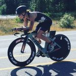 A woman biking. She is leaning low over her handle bars, dressed in black shorts, tank, and helmet. She is biking on a road with dry grass and some trees visible in the background.