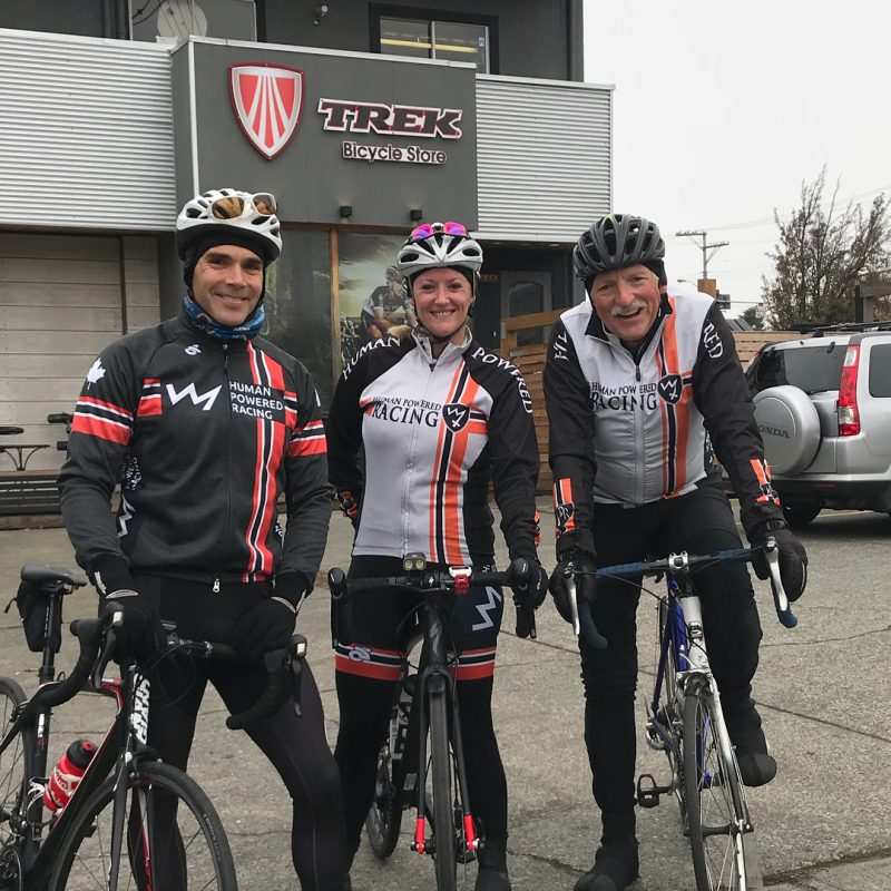 Three bikers in their gear with their bikes. Behind them on the left is a grey store with metal siding and the sign reads "Trek." It is a grey cloudy day.