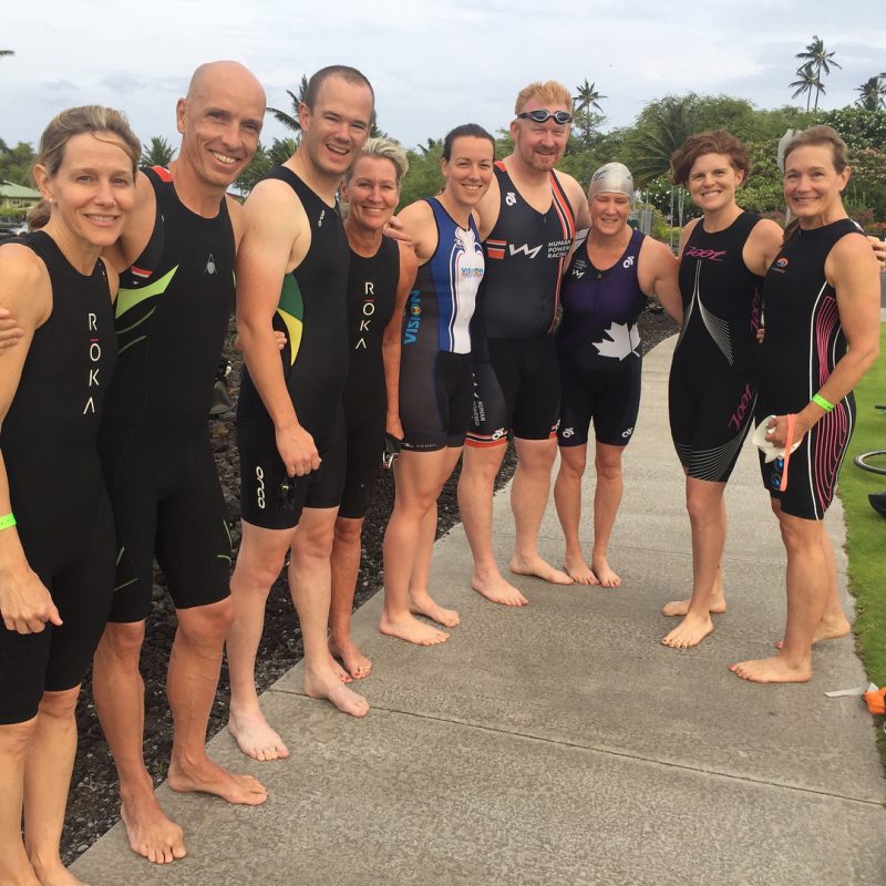 This is a photo of people standing in a J shape standing in a receeding line on the left and then curving around with more people in the centre and on the right. They are all in triathlon body suits and smiling. It is a cloudy day and there are palm trees in the background.