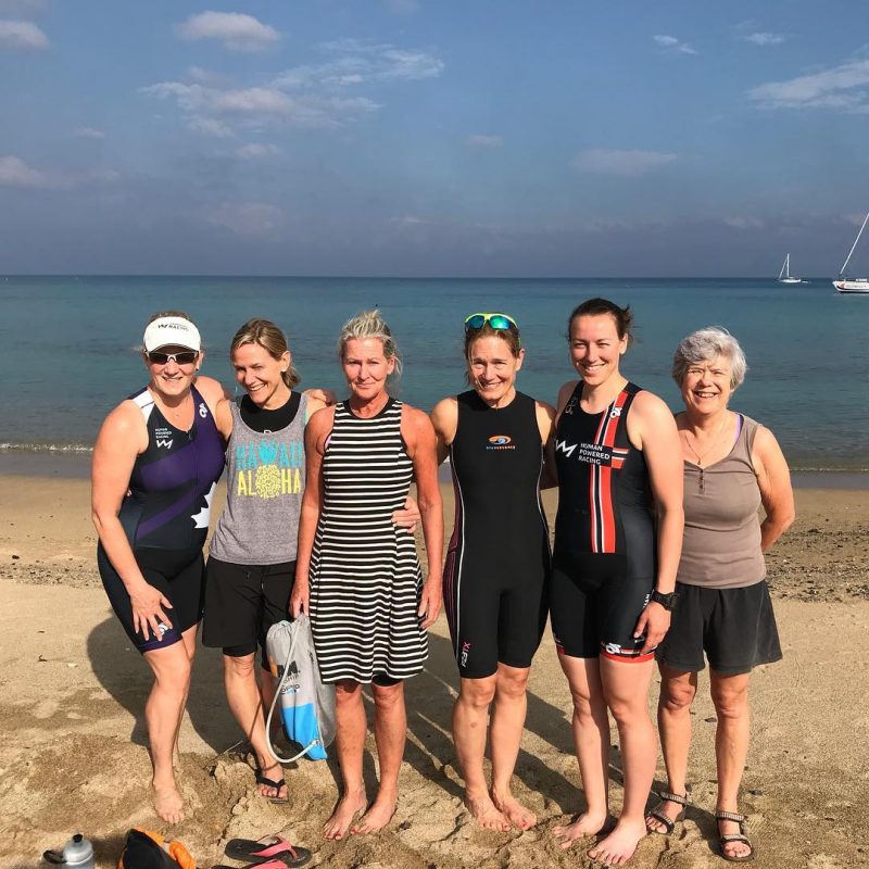 This is photo of six women standing on a beach with the ocean in the background on a sunny day. They are smiling at the camera. 