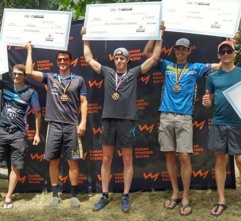 Five men standing in front of the black backdrop with the HPR logo on it, which is an orange line in the shape of a mountain range silhouette. They are holding large checks in the air.