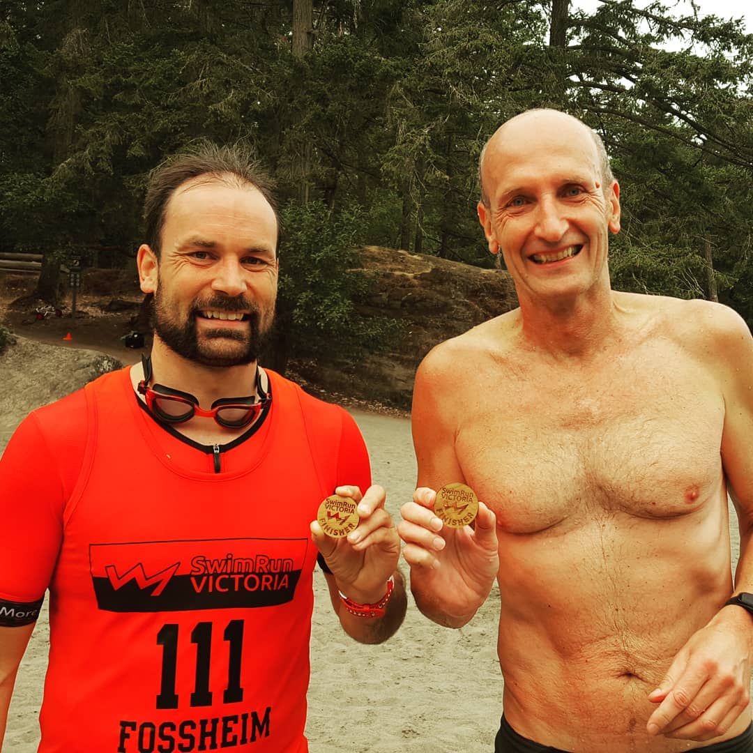 Two men standing shoulder to shoulder on a beach with evergreen trees in the back ground. Both are smiling and holding up wooden pendants or medals. The one on the left has a brown beard and orange tshirt. The one on the right is bald and shirtless. We see them from the waist up.