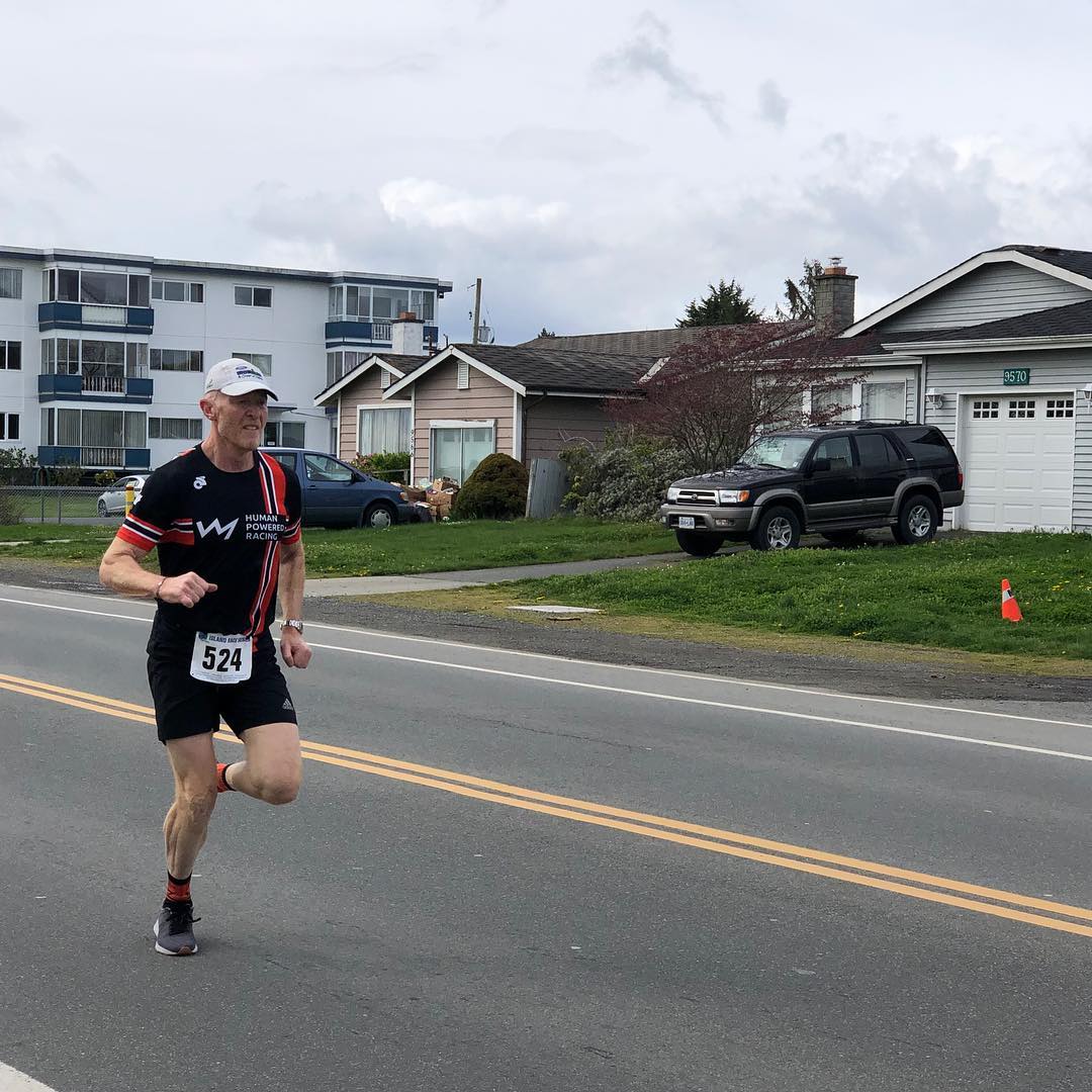 Rob in a black and red human powered racing shirt and black shorts running along a road.