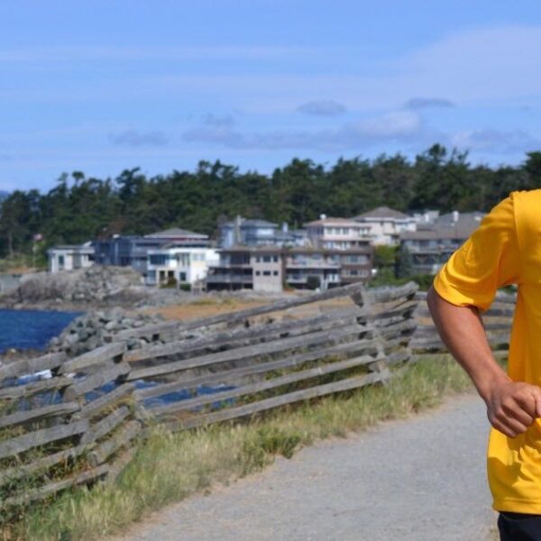 A man in a bright yellow shirt running and smiling is right of center. There is ocean in the background and his hair is blowing in the wind.