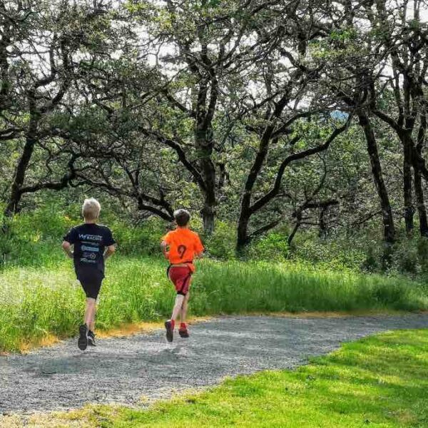 Two young people run away from the camera along a gravel trail with grass and trees on either side.