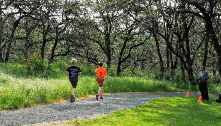 Two young people run away from the camera along a gravel trail with grass and trees on either side.