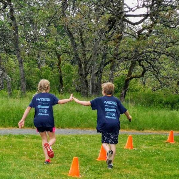 Two young people running away from the camera and high-fiving each other. They are in matching blue tshirts, and there are trees, grass, and orange pylons around them.
