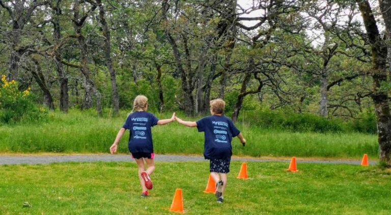 Two young people running away from the camera and high-fiving each other. They are in matching blue tshirts, and there are trees, grass, and orange pylons around them.