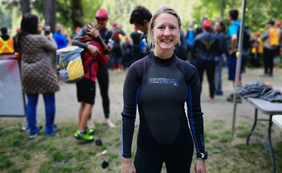 A woman in a wet suit with other athletes blurred out behind her smiling.
