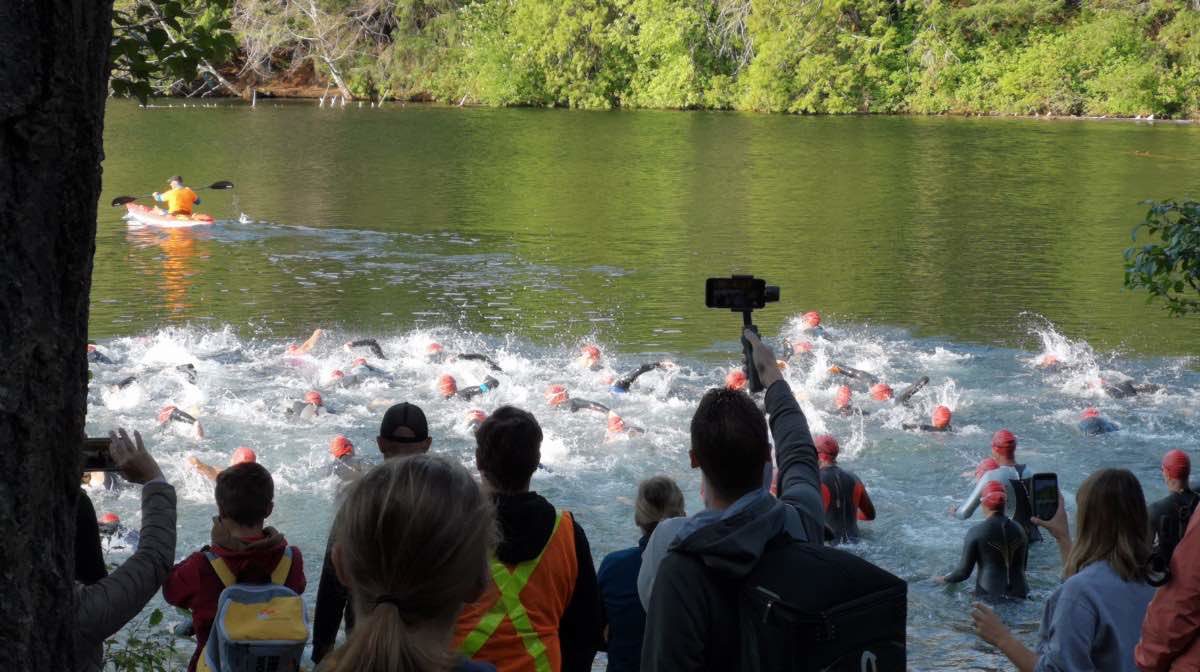 A crowd faces away from the camera towards a lake made turbulent by a crowd of racers starting the swim.