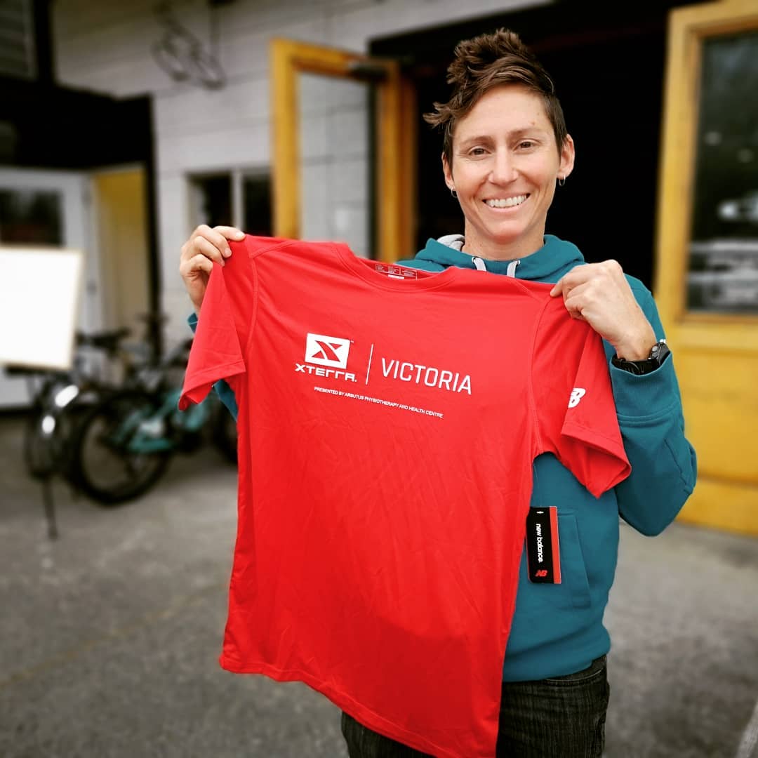 A woman holding up a red XTERRA tshirt and smiling outside of Trek Cycles.