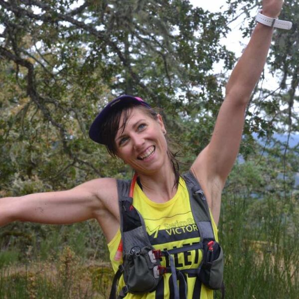 A woman smiles in a forest with her arms spread wide in celebration.