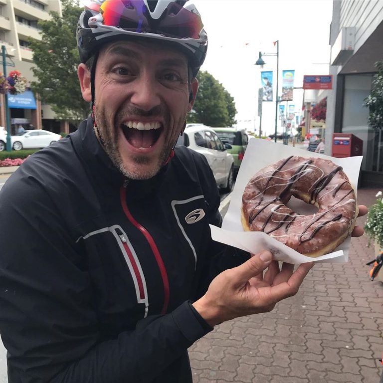 A male in a purple bike helmet and a black racing jacket holds up a donut on a napkin with chocolate and white sugar lines and grins with mouth open. Background is a city street