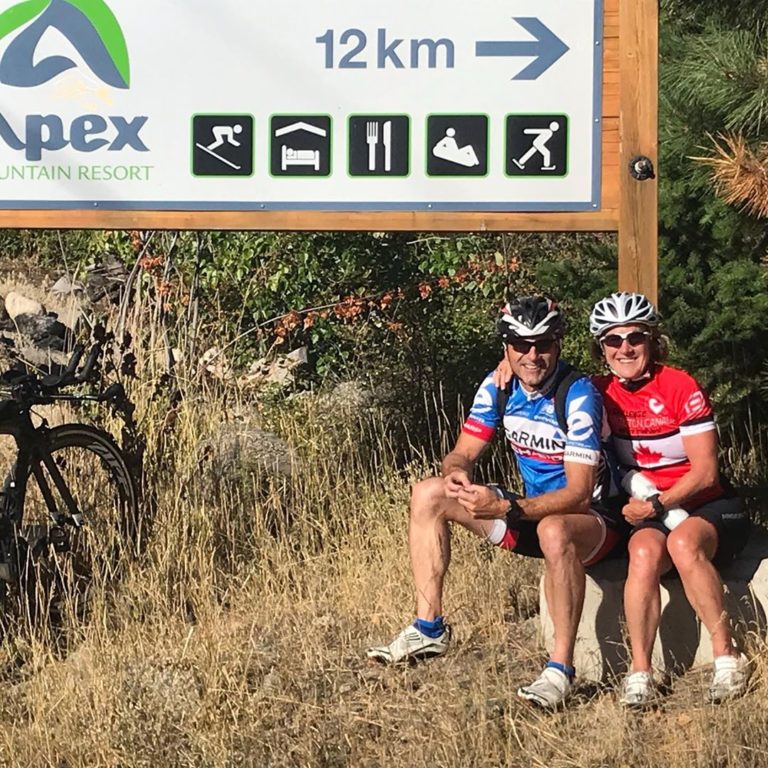 Two bikers in biking shorts and bright blue and red bike jerseys sit under a sign with helmets on. Bike is visible partially under the sign which says '12km' arrow to the right and signs for skiing, accomodation, eating and skating underneath