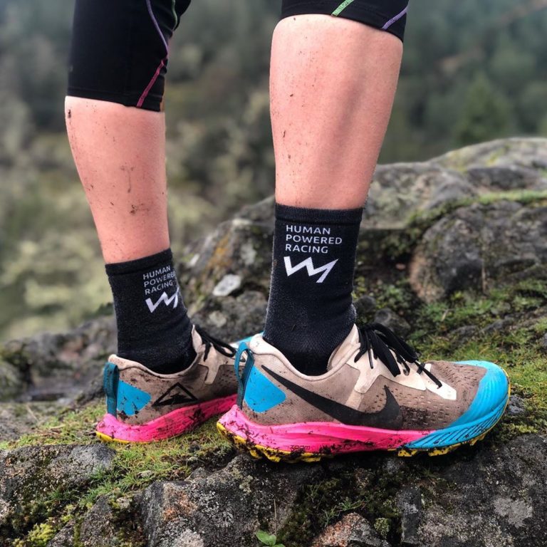 Ankle and feet of a runner in black socks and pink and blue running shoes visible standing on rock on a trail