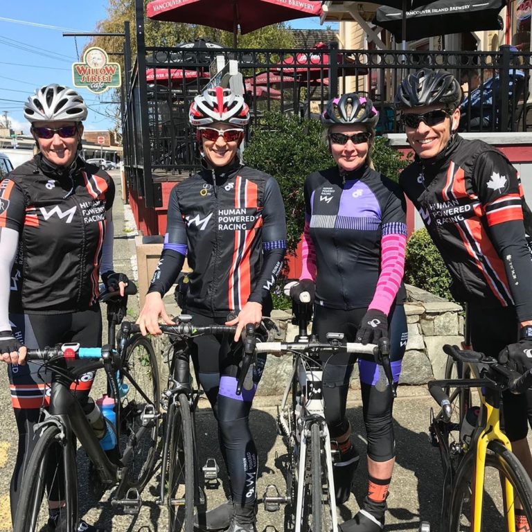 Four bikers in black biking gear and helmets pose with their bikes in front of a restaurant on a sidewalk in a city