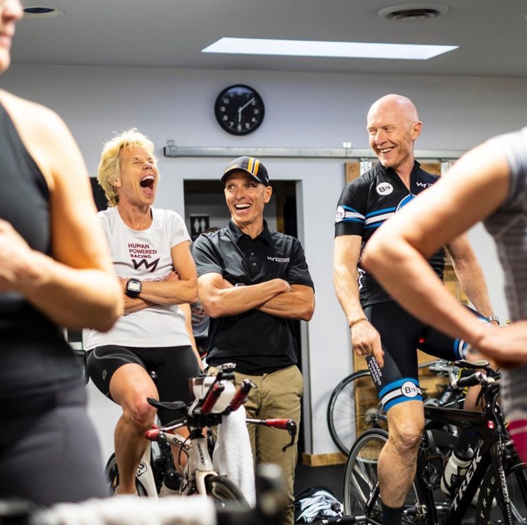 Two men on bikes are laughing exuberantly with mouths open, an elbow each of two other bikers visible and in the middle stands a coach for the stationary bikes.